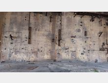 Eine alte, verwitterte Ziegelwand in einem verlassenen Gebäude mit sichtbaren Metallrohren, Halterungen und Spuren abgebauter Installationen; der Boden besteht aus beschädigten Betonplatten und Ziegeln.