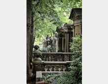 Historischer Friedhof mit einer Reihe von steinernen Mausoleen und dekorativen Geländern, umgeben von üppigem grünen Laub und überwachsenen Pflanzen. Die Szenerie wirkt schattig durch die dichten Baumkronen im Hintergrund.
