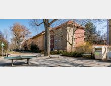 Ein Wohnviertel mit mehrstöckigen, farbigen Wohngebäuden und roten Ziegeldächern, flankiert von Bäumen und Gehwegen. Im Vordergrund stehen ein Tischtennistisch, Parkbänke und ein Altkleidercontainer.