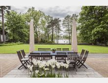 Große Gartenterrasse mit einem langen Holztisch und Stühlen, umgeben von gepflegtem Rasen und Bäumen. Im Hintergrund ist ein See zwischen den Bäumen sichtbar, mit Blumenbeeten und geschlossenen Sonnenschirmen im Vordergrund.
