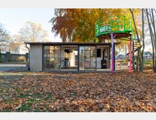 Kleines Gebäude mit großen Fenstern und einer offenen Front, umgeben von Herbstbäumen und gefallenen Blättern. Auf dem Dach befindet sich eine grün-gelbe Aussichtsplattform, die von pink-violetten Säulen gestützt wird.