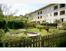 Spielplatz in einem begrünten Innenhof mit einem Sandkasten, einer Holzschaukel und einem Holzzaun. Im Hintergrund befindet sich ein mehrstöckiges Wohnhaus mit gelb umrandeten Fenstern und kleinen Gärten vor den Erdgeschosswohnungen.