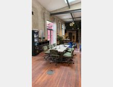 Großzügiger Arbeits- oder Besprechungsraum mit einem langen Tisch und mehreren grünen Bürostühlen auf einem Holzboden. Der Raum hat hohe Decken mit sichtbaren Metallträgern, große Fenster mit Vorhängen, eine Treppe, Pflanzen und dekorative Elemente wie eine Discokugel.