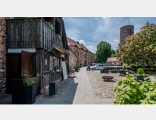 Historischer Innenhof mit gepflastertem Boden, flankiert von einem Fachwerkgebäude aus Holz und einem steinernen Bau mit roten Dächern; im Hintergrund ein runder Steinturm und ein blühender Kastanienbaum. Auf dem Platz stehen Bänke, Pflanzkübel und Autos.