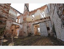 Innenansicht einer verfallenen Backsteinruine ohne Dach, mit freiliegenden Mauerresten, hohen Fensteröffnungen und einem teils erhaltenen Rundbogen. Im Vordergrund liegen überwucherte Ziegelböden und eine schief stehende Tür im offenen Durchgang.