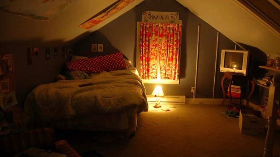 Cozy attic bedroom with sloped ceilings, a bed against the left wall, and a central window with patterned curtains lit by a floor lamp. A small TV on a stand and shelves with books and boxes line the right side of the carpeted room.