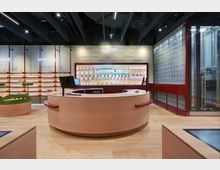 Innenraum einer modernen Apotheke mit einem runden Empfangstresen aus Holz im Vordergrund. Im Hintergrund befinden sich Regale mit Produkten sowie eine große Wand mit beleuchteten Fächern und Medikamenten hinter Glas.