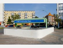 U-Bahnhof-Eingang "Innsbrucker Platz" mit einem modernen, grünen Schild und umliegender Bebauung aus Wohnhäusern und einem markanten Backsteingebäude mit rotem Dach; im Hintergrund sind Autos und Straßenszenen sichtbar. Der Boden vor dem Eingang besteht aus gepflastertem Gehweg.