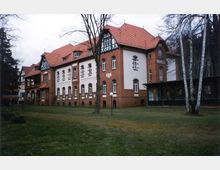 Backsteinbau mit roten Ziegeldächern im Stil der Gründerzeit, umgeben von einer Grünfläche mit vereinzelten Bäumen. Das Gebäude weist helle Putzflächen, verzierte Fensterrahmen und Fachwerkelemente auf.