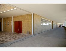 Überdachter Durchgang mit gelblichen Ziegelwänden, einer Reihe von großen Fenstern auf der rechten Seite und einer roten Doppeltür auf der linken Seite. Der Boden ist teilweise gefliest, teilweise aus Beton, und im Hintergrund ist ein Fahrzeug zu sehen.