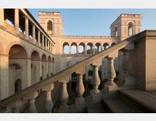 Historische Architektur mit einem offenen Hof, umgeben von beige- und rötlichfarbenen Arkaden aus Sandstein und Backstein. Im Vordergrund ist ein Treppenaufgang mit massiven Balustraden sichtbar, im Hintergrund verbinden Brückenbögen zwei höhere Gebäudeteile.