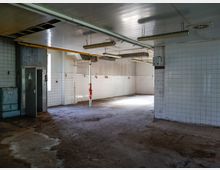 Leerer, stillgelegter Industrie- oder Küchenraum mit weiß gekachelten Wänden, Betonboden und einer abgehängten Decke mit Leuchtstoffröhren. Sichtbare Rohre, ein Metalltürbereich und wenige Fenster sorgen für spärliches Tageslicht.