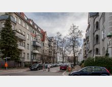 Straßenszene mit mehrstöckigen Wohnhäusern im Altbau-Stil auf beiden Seiten der Straße, roten Ziegeldächern und Balkonen. Geparkte Autos säumen die Straße, und zwei Fußgänger gehen auf dem Gehweg; kahle Bäume und ein leicht bewölkter Himmel im Hintergrund.