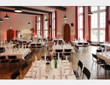 Ein großzügiger, heller Saal mit hohen Fenstern, weißen Wänden und Holzboden, eingerichtet für ein formelles Essen mit weiß gedeckten Tischen, schwarzen Stühlen und Tischdekoration wie Gläsern, Besteck und Stoffservietten. Im Hintergrund sind eine Bar, Holztüren und rote Vorhänge sichtbar, die die Fenster flankieren.