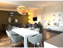 Moderne Wohnzimmer-Esszimmer-Kombination mit weißem Esstisch und grau gepolsterten Stühlen, einer L-förmigen grauen Couch, dunklem Holzboden und einer dekorativen Pendelleuchte. An einer Wand steht ein Sideboard mit künstlerischen Deko-Elementen, daneben eine große grüne Zimmerpflanze.