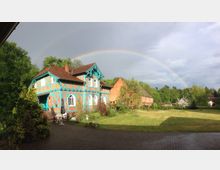 Ein traditionelles Fachwerkhaus mit auffälligen türkisfarbenen Balken vor einer weitläufigen grünen Wiese, umgeben von Bäumen. Im Hintergrund spannt sich ein Regenbogen über den hellen, teils bewölkten Himmel.