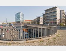 Straßenansicht mit mehrspuriger Autobahn, die durch einen Tunnel verläuft. Im Hintergrund sind ein modernes, rundes Bürogebäude mit Glasfassade, mehrere Wohnhäuser und ein entferntes Funkturmgebäude zu sehen; im Vordergrund ein Geländer und ein gepflasterter Gehweg.