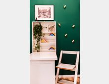 Ein kleiner, moderner Innenraum mit einer weißen Ablage, einem Regal mit Pflanzen, Büchern und Dekorationen vor einer dunkelgrünen Wand. An der Wand hängen Haken, und ein schwarz-weißes gerahmtes Bild ist darüber angebracht; daneben steht ein minimalistischer weißer Stuhl mit Holzdetails.