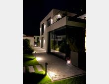 Moderne Wohnhausfassade bei Nacht mit großen Glasflächen und weißem Putz. Der gepflasterte Weg vor dem Haus ist durch kleine, eingelassene Bodenlampen beleuchtet, und der angrenzende Rasen ist gepflegt.