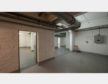 Kellerraum mit weißen Betonwänden, grauem Boden und sichtbaren Belüftungsrohren an der Decke. Ein geöffneter Durchgang führt in einen angrenzenden Raum, während an der Wand ein Elektrokasten und daneben ein einsamer Stuhl zu sehen sind.