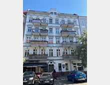 Fünfgeschossiges Wohnhaus im Altbaustil mit weißen Fassaden und mehreren Balkonen, im Erdgeschoss befinden sich eine Pizzeria und ein Friseursalon. Davor stehen Autos geparkt, und einige Bäume rahmen die Szene ein.