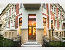 Fassade eines historischen Gebäudes mit rotem Klinker und weißen Stuckelementen, zentral eine doppelflügelige Holztür mit Glasfenstern und darüber leuchtender Schriftzug. Vorgarten mit schmiedeeisernem Zaun und Treppenstufen, die zum Eingang führen.