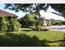 Steinhaus mit rotem Ziegeldach, umgeben von Pflanzen und Rankgewächsen, in einem Garten mit Rasenfläche und Blumenbeeten; im Vordergrund Schatten von Bäumen, rechts ein gepflasterter Bereich.