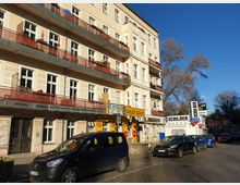 Mehrstöckiges, beiges Wohn- und Geschäftsgebäude mit Balkonen, einem Schilderladen im Erdgeschoss und parkenden Autos entlang der Straße. Im Hintergrund sind Bäume und ein blauer Himmel zu sehen.