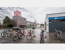 Straßenszene mit einem Einkaufszentrum im Hintergrund, erkennbar an einem "Kaufland"-Schild, einer U-Bahn-Station der Linie U Britz-Süd rechts und mehreren Fahrrädern, die an Fahrradständern entlang des Gehwegs abgestellt sind. Der Himmel ist bewölkt, und eine Straße mit fahrenden Autos verläuft durch das Bild.