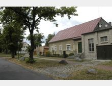 Straßenszene in einem Dorf mit einer gepflasterten Straße und Bäumen am Rand. Rechts steht ein beige-graues Wohnhaus mit einem roten Ziegeldach, Fenstern und einer grünen Tür, daneben ein älteres Gebäude aus heller Ziegelsteinfassade.