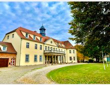 Historisches Gebäude mit gelber Fassade, roten Ziegeldächern und einem kleinen Turm in der Mitte, umgeben von einem Kiesweg und einer Rasenfläche. Rechts im Bild stehen Bäume, teilweise mit Herbstlaub.