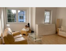 Wohnzimmer mit hellem Holzfußboden, einer gelben Ledercouch, einem Glastisch und einem zweiten kleineren Sofa. Im Hintergrund sind große Fenster mit Blick auf Bäume und angrenzende Gebäude zu sehen, neben denen eine aufgestellte Matratze und eine Stehlampe stehen.