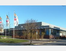 Modernes Gebäude mit flachem Dach und großen Fenstern, umgeben von einem kleinen Park mit Bäumen und Wegen. Links im Bild sind mehrere bunte Fahnen zu sehen, neben einer Grünfläche.