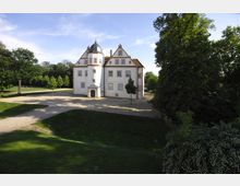 Historisches Gebäude mit weißer Fassade, symmetrischen Fenstern und einem kleinen Turm mit zwiebelförmiger Kuppel, umgeben von einem weitläufigen Garten mit Rasenflächen, Bäumen und gepflasterten Wegen.