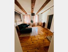 Helles Wohnzimmer mit breiten Holzdielen und sichtbaren Deckenbalken, zwei Fenstern mit hellen Vorhängen und einer dunklen Pendelleuchte. Dunkelgrünes Sofa mit Couchtisch auf einem Teppich sowie eine Kommode an der rechten Wand.