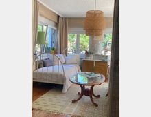 Schlafzimmer mit einem weißen Metallbett, daneben ein Holzschrank mit Dekorationen. Ein runder Glastisch steht auf einem Teppich, über dem eine hängende Lampe aus Korbgeflecht hängt. Große Fenster mit beigen Vorhängen bieten Blick auf einen Garten.