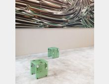 Minimalistischer Innenraum mit hellem Betonboden, einer beigen Wand und einer großformatigen Wandkunst mit wellenförmigen, metallischen Mustern. Im Vordergrund stehen zwei kleine, grünliche Sitzhocker aus gitterförmigem Kunststoff.