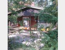 Holzhütte im Wald, umgeben von dichten Nadelbäumen. Die Hütte hat eine kleine Veranda mit einem Geländer und gelben Gartenstühlen; im Hintergrund ist ein rotes Ziegeldach sichtbar.