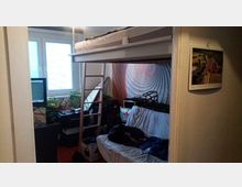 Ein kleines Zimmer mit Hochbett und Leiter, darunter ein Sofa mit einer weißen Decke. An der Wand ist eine spiralförmige Dekoration, auf der Fensterbank stehen Pflanzen, und ein Schreibtisch mit Computerbildschirm befindet sich neben dem Fenster.