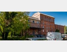 Backstein-Gebäude im Stil eines historischen Lagerhauses mit mehreren Etagen und schmalen Fenstern, umgeben von Bäumen und geparkten Fahrzeugen im Vordergrund. Ein Schild am Dach weist auf eine mögliche industrielle Nutzung hin.