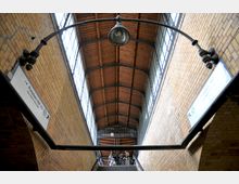 Bahnhofshalle mit gelben Klinkerwänden, einer hohen Holzdecke und großen Fenstern entlang der Seiten. Im Vordergrund eine Aufhängung mit alter Lampe, im Hintergrund eine Treppe mit Personen und Fahrrädern, die zu einer Glastür mit einem Uhrensymbol führt.