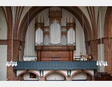 Innenansicht einer Kirche mit einer großen, verzierten Orgel aus Holz und Pfeifen im neugotischen Stil, die auf einer Empore über Bögen aus roten Ziegeln steht. Im Vordergrund hängen mehrere schlichte weiße Kronleuchter von der Decke.