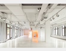 Großer, lichtdurchfluteter Veranstaltungsraum mit weißen Wänden und hellem Boden. Die Decke ist mit sichtbaren Lüftungsrohren, Schienenleuchten und technischen Einrichtungen ausgestattet, während die großen Fenster mit weißen Vorhängen versehen sind.