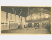 Historische Turnhalle mit Holzbalkenkonstruktion und hoher Decke, ausgestattet mit Kletterseilen, Turnringen, Sprossenwänden, Turnpferden und Leitern. Im Hintergrund befinden sich Türen und Fenster sowie eine erhöhte Empore mit Geländer.