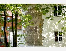 Fassade eines Gebäudes mit weißem Putz, auf dem ein kunstvolles Sonnenuhr-Motiv mit Sonnen- und Monddarstellung sowie dekorativen Figuren zu sehen ist, umgeben von Laubbäumen im Vordergrund. Im Hintergrund ein weiteres Gebäude mit rotem Ziegelmauerwerk und Fensterläden.