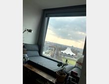 Ein Fenster mit Sitzbereich im Inneren eines Raums blickt auf eine Stadtlandschaft mit dem markanten Zeltdach des Berliner Tempodroms. Im Vordergrund sind eine kleine Topfpflanze, ein graues Sitzpolster und Bücher auf einem Holzregal sichtbar.