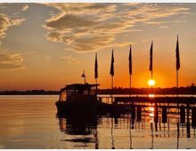 Sonnenuntergang an einem ruhigen See mit einem Bootsanleger, auf dem ein Boot und mehrere Fahnenmast-Silhouetten stehen. Im Hintergrund reflektiert die tief stehende Sonne orangefarbenes Licht auf der Wasseroberfläche.