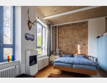 Schlafzimmer mit modernem und rustikalem Design, bestehend aus einer freiliegenden Ziegelwand, einem Holzbett mit blauer Bettwäsche, einem Parkettboden sowie großen Fenstern mit blauen Rahmen. An der weiß gestrichenen Wand hängen ein Geweih und dekorative Kunstwerke, daneben befindet sich ein kleiner Kamin.