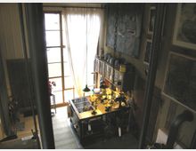 Werkstatt in einem rustikalen Raum mit Holzboden und Holzwänden. Ein großer Arbeitstisch ist mit Werkzeugen, Materialien und Beleuchtung ausgestattet, daneben hängen Regale mit Dosen und Behältern, während durch hohe Fenster mit Vorhängen natürliches Licht einfällt.