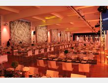 Großer Veranstaltungsraum mit langen, gedeckten Tischen und weißen Stühlen, beleuchtet von warmem orangefarbenem Licht. Die Wände sind mit großflächigen, abstrakt-geometrischen Wandbildern verziert, und an der Decke hängen Beleuchtungselemente.